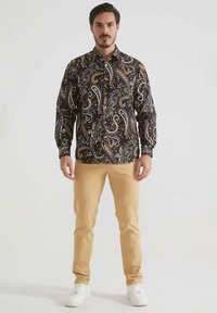 Chemise à manches longues en paisley dans des tons sombres avec des motifs complexes, associée à un pantalon beige et des baskets blanches. Tissu doux, coupe régulière.
