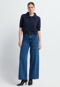 Marineblauer, kurzer Cardigan mit großem Kragen und kurzen Ärmeln, kombiniert mit hochtaillierten, weit geschnittenen blauen Jeans. Schwarze, spitz zulaufende Absatzschuhe.