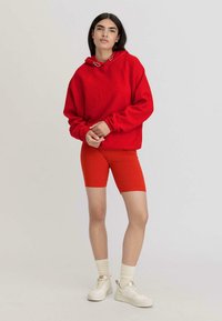 Felpa rossa oversize con cordini e occhielli in metallo, abbinata a pantaloni corti da ciclismo rossi elasticizzati. Il modello indossa sneakers bianche e calze color crema.