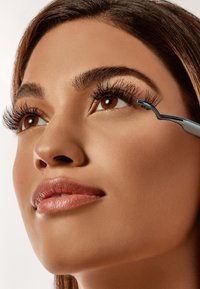 Primo piano di una persona con lunghe e voluminose ciglia, che tiene un piegaciglia argento vicino all'occhio, pelle levigata e un colore di labbra neutro.