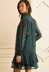 Rochie cu mânecă lungă de un teal închis, cu modele abstracte colorate, guler și tiv volanat. Fabricată din material transparent, are manșete strâmte.