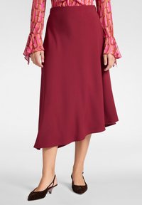 Falda A-line burdeos hecha de tela suave, con un dobladillo asimétrico. Combinada con zapatos slingback marrones de textura.