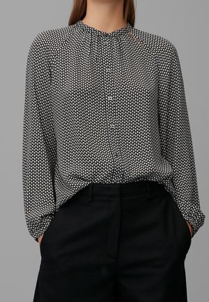 Vrouw draagt een zwart-wit gebloemde blouse met knopen, ingestopt in hoge zwarte broek, handen in de zakken, staand tegen een grijze achtergrond.