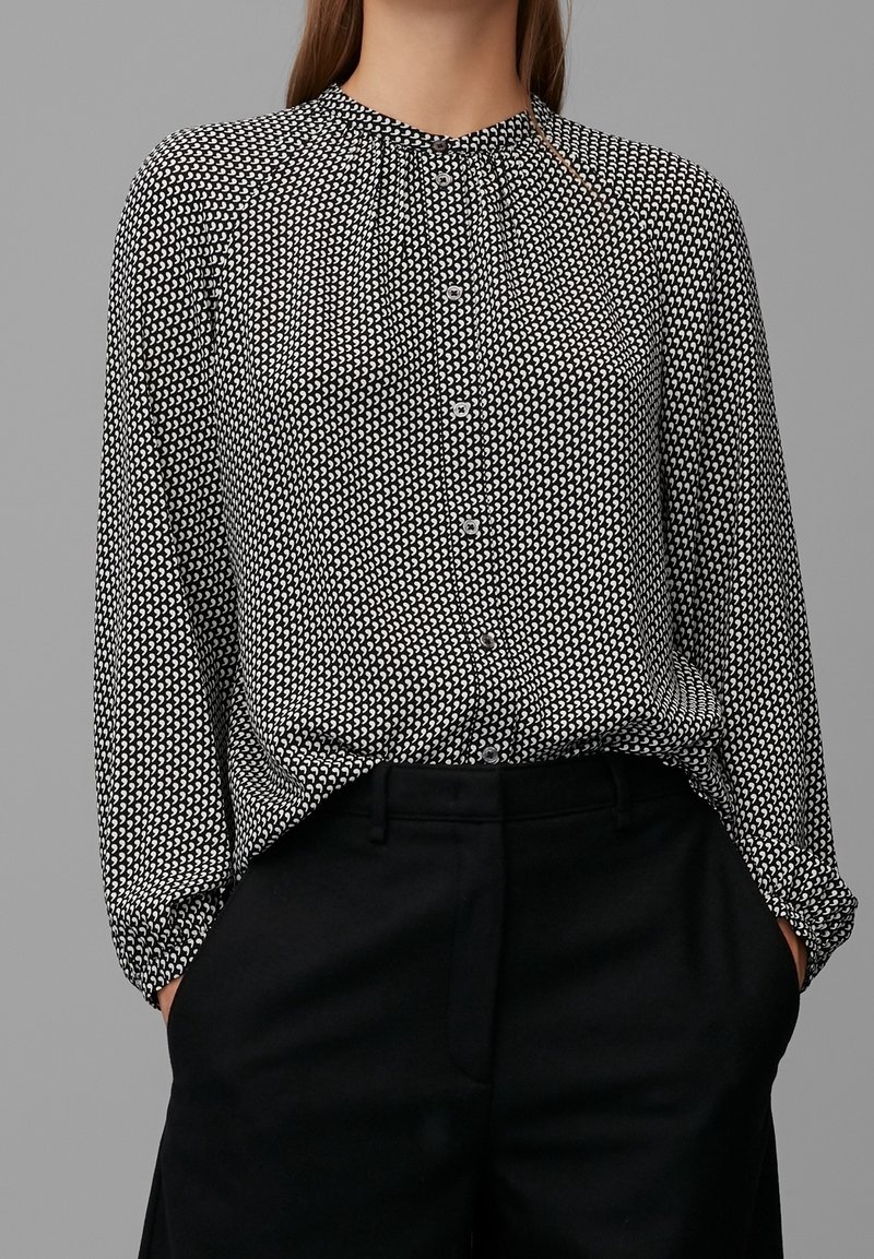 Femme portant un chemisier à motifs noir et blanc boutonné, rentré dans un pantalon noir taille haute, mains dans les poches, debout devant un fond gris.