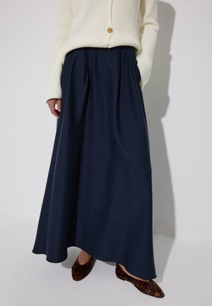 Naine, kes kannab kreemjat kootud kampsunit, tumelõvi pikka seelikut taskutega ja leopardimustrilisi madalaid kingi, seisab ühe käega taskus.