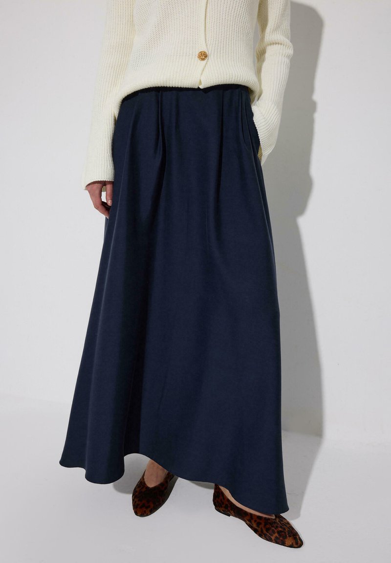 Donna che indossa un maglione lavorato a maglia color crema, gonna lunga blu navy con tasche e ballerine con stampa leopardata, in piedi con una mano in tasca.