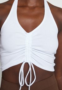 Gros plan d'une personne portant un crop top blanc froncé avec un col en V et un cordon de serrage à l'avant, associé à un pantalon côtelé marron.