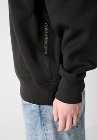 Hanorac negru cu manșete și tiv din tricot, având un eticheta logo în gri pe lateral. Purtați cu blugi din denim albastru deschis.
