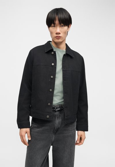 Νεαρός άνδρας με μαύρο πουκάμισο με κουμπιά πάνω από ανοιχτό πράσινο πουκάμισο και ξεθωριασμένο μαύρο τζιν, στέκεται μπροστά σε απλό λευκό φόντο.