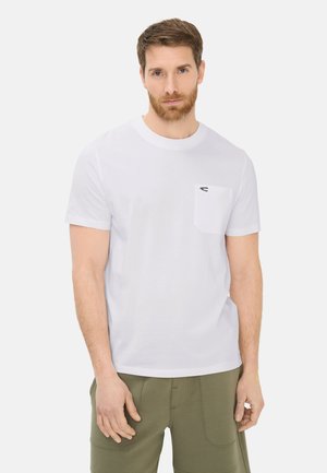 Mann mit kurzem braunem Haar und Bart, der ein weißes T-Shirt mit kurzen Ärmeln und Brusttasche sowie olivgrüne Hose trägt, vor einem einfarbigen Hintergrund stehend.