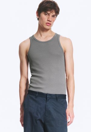 Jonge man die een aansluitende grijze mouwloze geribde tanktop en donkerblauwe broek draagt, staand tegen een effen lichte achtergrond.