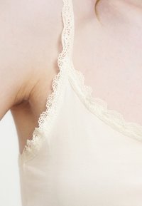 Gros plan d'une personne portant un caraco couleur crème avec une délicate bordure en dentelle le long de l'encolure et de la bretelle d'épaule.