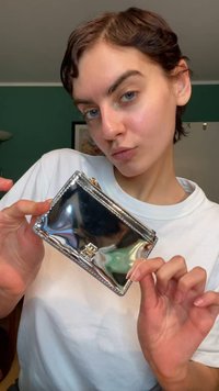 Metallic silver wallet with a smooth texture, zip closure, and gold hardware detail featuring a logo on the front. Compact rectangular shape.