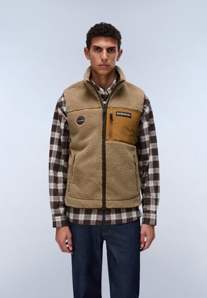 Mand iført beige fleecevest med brun brystlomme over brun og hvid ternet skjorte og mørkeblå jeans, stående foran ensfarvet grå baggrund.