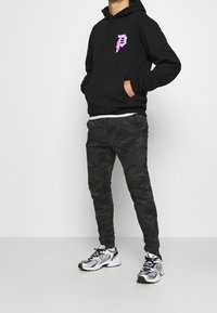 Černá mikina s kapucí s růžovým logem, doplněná úzkými černými maskáčovými tepláky a stříbrnými sportovními botami.