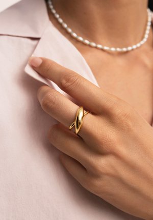 Goldring mit einer glatten, länglichen Form, getragen an einer Hand mit heller Haut, ausgestattet mit einem rosa Hemd mit Kragen und einer Perlenkette.