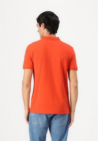 Camisa polo naranja con cuello clásico y mangas cortas, fabricada en tejido suave. La parte trasera presenta un dobladillo recto. Combinada con jeans azules.