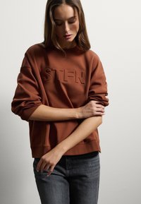 Felpa marrone con logo "STFN" in rilievo, scollo rotondo, spalle scese e polsini a coste, abbinata a jeans grigio scuro.