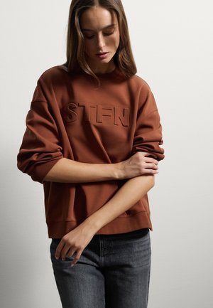 Felpa marrone con logo "STFN" in rilievo, scollo rotondo, spalle scese e polsini a coste, abbinata a jeans grigio scuro.