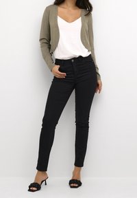 Schwarze, schmal geschnittene Jeans kombiniert mit einer verkürzten beigen Strickjacke über einem weißen V-Ausschnitt-Shirt. Das Modell trägt schwarze, high-heeled Sandalen mit einem geflochtenen Design.