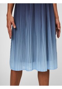 Plissé midi rok met een verloop van donkerblauw naar lichtblauw, gedragen door een persoon met een gebruinde huid, met ontspannen handen naast het lichaam.