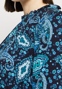 Navy blue blouse featuring intricate turquoise paisley and floral patterns, with a ruffled neckline and soft fabric texture.