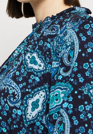 Blouse bleu marine présentant des motifs complexes de paisley et de fleurs turquoise, avec un col volanté et une texture de tissu douce.