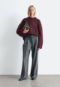 Burgunderfarbener, flauschiger Pullover, weite graue Hosen und elegantes schwarzes Schuhwerk. Hält eine strukturierte braune Tasche. Schlichter Hintergrund in neutralen Farben.