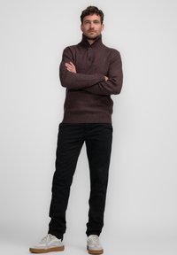 Suéter de punto marrón con un cuello alto, que presenta dos botones, combinado con pantalones negros y zapatillas de color claro. Diseño sencillo, tela texturizada.