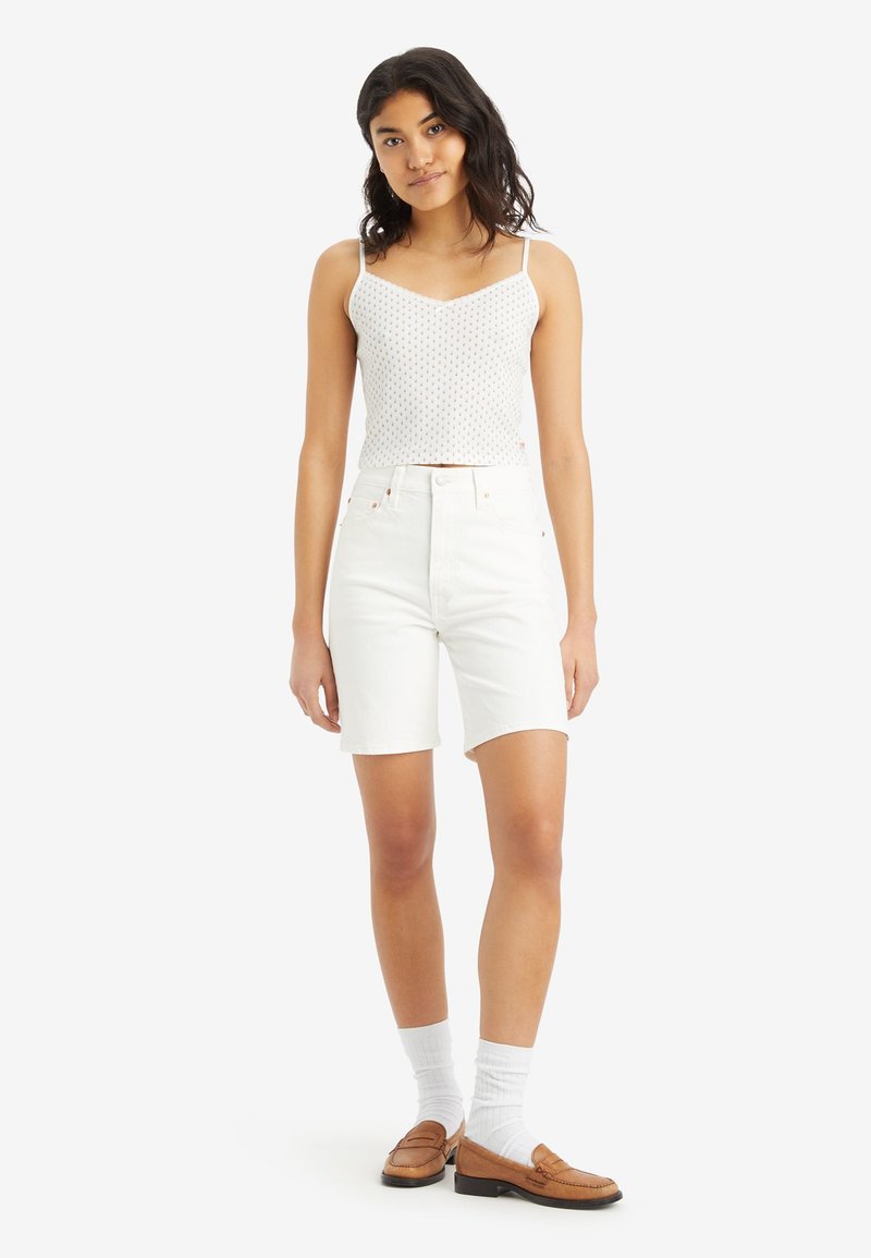 Top corto bianco senza maniche con piccoli punti colorati, abbinato a pantaloni corti di denim bianchi fino al ginocchio e mocassini marroni con calze bianche.