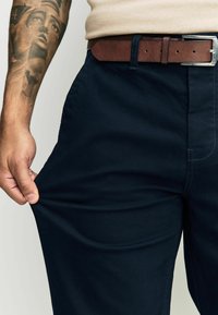 Sötétkék nadrág sima textúrával és egyenes szabással, barna bőrövvel és látható varrással az oldals seams-eken.