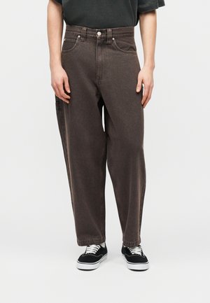 Άτομο που φοράει άνετα σκούρα καφέ τζιν και μαύρα αθλητικά παπούτσια με λευκές σόλες και κορδόνια, στέκεται μπροστά σε απλό ανοιχτό φόντο.