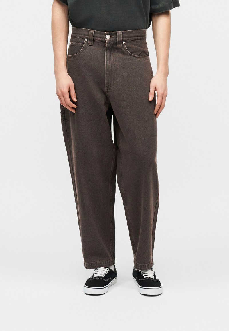 Osoba oblečená v voľných tmavohnedých džínsoch a čiernych teniskách s bielymi podrážkami a tkaničkami, stojaca na jednoduchom svetlom pozadí.