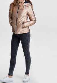 Persoon in glanzende beige pufferjack met rits, donkere skinny jeans en witte sneakers, staand met handen in de zakken van de jas.