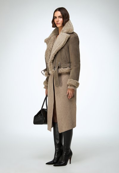 Veste marron doublée en shearling avec une taille ceinturée, associée à une jupe côtelée beige et de hautes bottes noires à talons aiguilles, tenant un sac à main noir.
