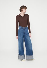 Mujer con una camiseta de manga larga de canalé marrón y pantalones vaqueros anchos de mezclilla azul con los puños doblados, de pie frente a un fondo claro y liso.