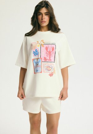 Woman wearing a white oversized t-shirt with colorful seashell, lobster, and sardine can illustrations, paired with white shorts.