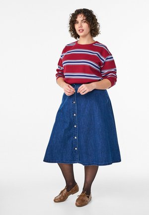 Ženska s kodrastimi lasmi, oblečena v rdečo majico z dolgimi rokavi s črtami, moder jeans midi krilo z gumbi, črne nogavice in rjave čevlje, stoji vzravnano.