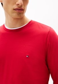 Rode blouse met lange mouwen van gladde stof, met een witte ronde hals en een klein geborduurd logo op de borst.