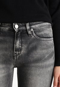 Black denim jeans with a faded, textured finish, five-pocket design, metal button closure, and a slim fit.