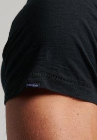 T-shirt nera realizzata in tessuto morbido con una texture liscia. Presenta un orlo cucito e una piccola etichetta con la scritta "SUPERDRY" in blu.