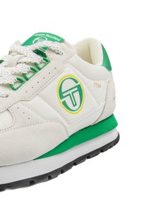 White sneaker with green accents, suede and mesh materials, round logo in green and yellow, featuring a cushioned sole and perforated details.