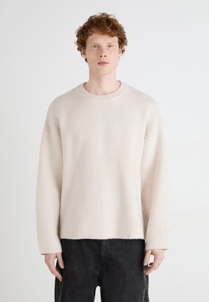Lys beige strikket genser med rund hals, nedfalne skuldre og lange ermer, kombinert med mørke, strukturerte bukser.