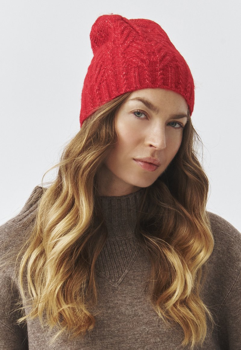 Red knitted beanie with a textured cable pattern, featuring a tapered crown and a subtle roll at the hem.