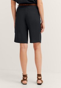 Dunkelgraue maßgeschneiderte Shorts mit einer schlanken Passform, strukturiertem Material und zwei Gesäßtaschen, kombiniert mit braunen Wildleder-Sandalen mit gestapeltem Absatz.
