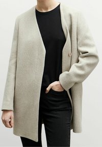 Ljusgrå ullblandad kappa med öppen front, knappstängning och långa ärmar, bärs över en svart topp och svarta byxor.