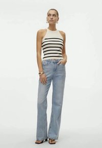 Kvinna i en beige ärmlös randig topp, ljusblå högmidjade jeans, hoopörhängen, armband och svarta sandaler med öppen tå.