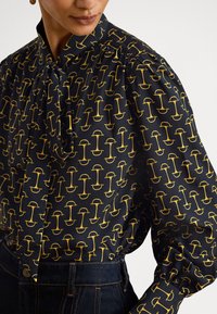 Crna bluza s vrpcom oko vrata, ukrašena ponavljajućim žutim grafičkim uzorkom. Duge nabrane rukave s elastičnim manšetama. Nošena uz tamni traper.