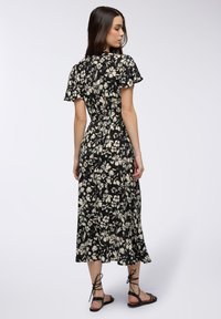 Abito floreale nero con maniche corte, vita aderente e gonna a forma A. Presenta motivi floreali bianchi e una texture fluida. Sandali neri con cinturini.