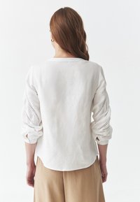 Valge linane pluus pikkade varrukatega, millel on tikitud silmade detailid ja ümardatud ääred, näidates seljast lõdva istuvust.